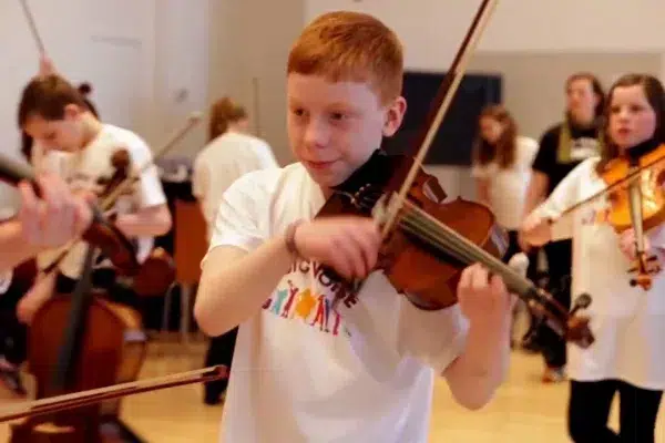 young-boy-with-short-red-hair-white-tishirt-playing-the-violin-with-other-young-people-in-a-community-hall